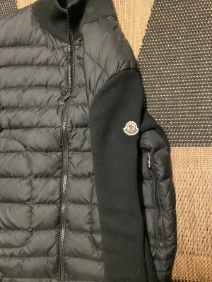 Svart cardigan jacka från Moncler - Snygg svart cardigan jacka från Moncler med quiltad framsida och stickade ärmar.  Storlek M men passar S det ända negativa är att monclear märket på sidan har lite defekt. Jackan har dragkedja framtill, hög krage och en Moncler-logga på ena ärmen. Perfekt för dig som vill ha en stilren och exklusiv look.
