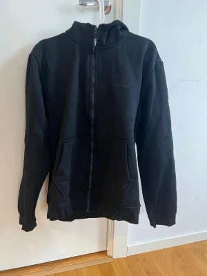 Svart hoodie från C.P. Company - Svart hoodie med dragkedja från C.P. Company. Klassisk design med två stora fickor framtill och diskret logga på bröstet. Huvan har inbyggda goggles som ger en unik och streetinspirerad look. Tillverkad i mjukt bomullsmaterial. Liten i storleken passar M och L den är helt ny oanvänd 