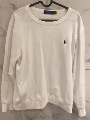 Vit sweatshirt från Polo Ralph Lauren - Klassisk vit sweatshirt från Polo Ralph Lauren med rund halsringning. Tröjan har långa ärmar, ribbade muddar och är gjord i mjukt bomullsmaterial. Perfekt för en clean och stilren look.