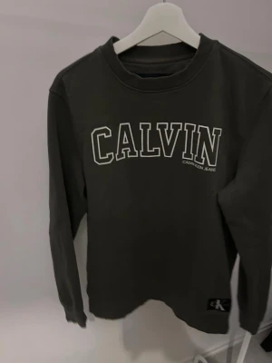 Olivgrön Calvin Klein Jeans sweatshirt - Snygg olivgrön sweatshirt från Calvin Klein Jeans med vit logotyp framtill. Tröjan har rund halsringning och ribbade muddar vid ärmslut och nederkant. Perfekt för en avslappnad och trendig stil.