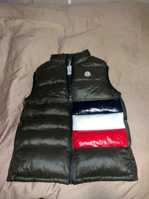 Moncler dunväst i olivgrönt - Snygg olivgrön dunväst från Moncler med hög krage och dragkedja framtill. Västen har en cool detalj med färgblock i marinblått, vitt och rött på ena sidan samt Moncler-logga på bröstet. Perfekt för lager på lager och streetwear-vibe.