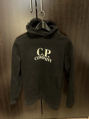 Svart hoodie från C.P. Company - Snygg svart hoodie från C.P. Company med stor vit logga på bröstet och klassiska goggles i huvan. Hoodien har känguruficka framtill och är gjord i mjukt bomullsmaterial. Perfekt för dig som gillar streetwear och vill ha något stilrent men ändå unikt.
