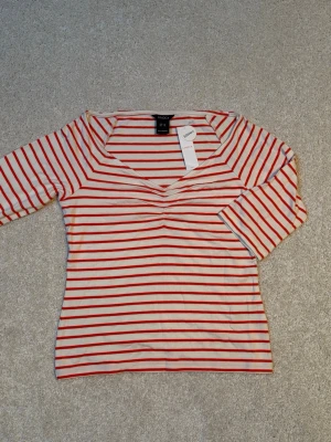Randig topp från LINDEX med v-ringning, ny med prislapp. - Säljer en vit och röd randig topp från LINDEX med v-ringning och trekvartsärm. Toppen har en smickrande passform och tunna röda ränder över hela plagget. Perfekt för dig som gillar en enkel men snygg stil. Helt ny och oanvänd, prislapp kvar. Nypris 199kr