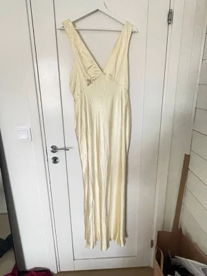 Ljusgul långklänning med v-ringning - Säljer en elegant ljus långklänning med djup v-ringning och breda axelband. Klänningen har en mjuk, glansig yta och följsam passform som ger en snygg siluett. Perfekt för dig som vill ha en stilren och enkel look till balen