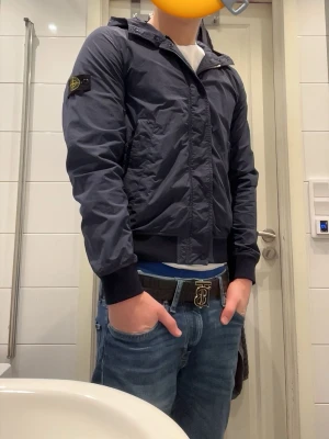 Mörkblå vindjacka från Stone Island - Snygg och stilren mörkblå vindjacka från Stone Island med ikonisk patch på vänster ärm. Jackan har huva, dragkedja framtill och ribbade muddar vid ärmslut och midja. Perfekt för dig som vill ha en snygg och funktionell jacka till vardags. Jag är 178 och den är lite liten för mig.