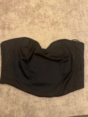 Svart axelbandslös korsettopp - Snygg svart korsettopp med axelbandslös design och markerad byst. Toppen har en dragkedja i ryggen för enkel på- och avtagning. Perfekt att styla med höga jeans eller kjol för en trendig look.