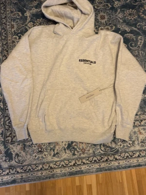 Grå Essentials Fear of God hoodie - Snygg ljusgrå hoodie från Fear of God Essentials med svart logga på bröstet. Klassisk passform med huva och känguruficka framtill. Perfekt för en avslappnad och trendig streetwear-look.