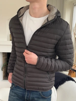 Svart pufferjacka med huva Colmar - Snygg svart pufferjacka med huva och dragkedja framtill. Jackan har quiltad design och två sidofickor. Perfekt för kyliga dagar och ger en modern look. Passformen är normal och jackan är tillverkad i lätt syntetmaterial.