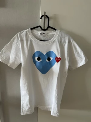 Vit Comme des Garçons Play t-shirt - Säljer en vit t-shirt från Comme des Garçons Play med det ikoniska blå hjärtat med ögon tryckt på bröstet och ett litet rött hjärta broderat bredvid. T-shirten har rund hals och korta ärmar. Tillverkad i mjuk bomull, perfekt för en clean och trendig look.