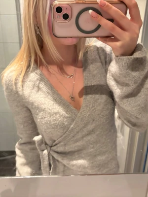 Grå omlottstickad tröja - Mysig grå stickad tröja med omlottmodell och knyt i sidan. Tröjan har v-ringning och långa utsvängda ärmar.🤍