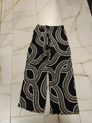Svarta vida byxor med vit swirl - Säljer ett par svarta byxor med vitt swirl-mönster. Byxorna har en loose och wide passform med hög midja och croppad längd. Perfekta för dig som vill sticka ut med ett unikt mönster. Materialet känns mjukt och skönt, troligtvis bomull eller polyester.