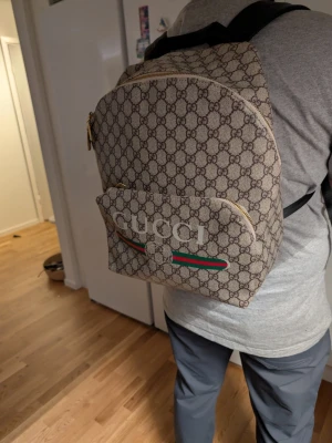 Ryggsäck med GG-mönster - Säljer en snygg ryggsäck i beige med klassiskt GG-mönster och gröna/röda ränder på fickan. Väskan har guldfärgade dragkedjor och är tillverkad i canvas med skinn-detaljer. Perfekt för dig som vill sticka ut med en ikonisk design. Akopia