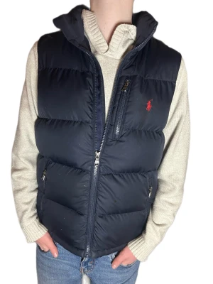 Mörkblå dunväst från Polo Ralph Lauren - Snygg mörkblå dunväst från Polo Ralph Lauren med hög krage och dragkedja framtill. Västen har två sidofickor med dragkedja och en bröstficka samt den klassiska röda loggan broderad på bröstet. Defekter asså fläckar syns enligt mig bara i stark ljus men inget man tänker på🙂 Modellen är 155cm. Ralph Lauren västen är i M i barn storlek. Skickar snabbt