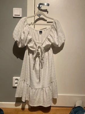 Vit kort klänning från Gina Tricot - Supersöt vit kort klänning från Gina Tricot med puffärmar och knytning framtill. Klänningen har en volang nedtill och är gjord i ett strukturerat bomullsliknande material. Perfekt för sommarens alla tillfällen!Enbart andvänd på en skolavslutning!