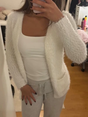 Vit fluffig kofta med fickor - Mysig och fluffig vit kofta med långa ärmar och två praktiska fickor framtill. Koftan har öppen front och är tillverkad i ett mjukt, lurvigt material som känns superbekvämt. Perfekt att slänga på sig över en topp för en avslappnad look.