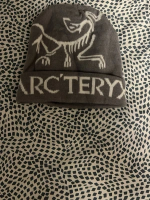 Grå mössa från Arc'teryx - Snygg grå mössa från Arc'teryx med vit logga och text framtill. Tillverkad i mjukt, stretchigt material som sitter skönt på huvudet. Perfekt för kalla dagar och outdoor-aktiviteter. Klassisk passform och stilren design.