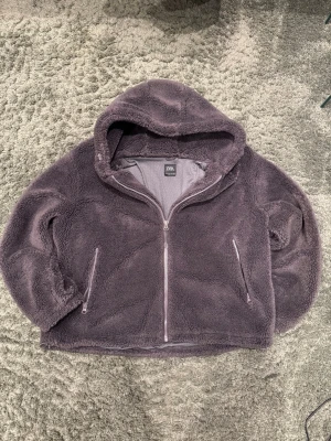 Fleecejacka med huva från Zara - Mysig mörklila teddyjacka från Zara med huva och dragkedja framtill. Jackan har två fickor med dragkedja och är tillverkad i mjuk syntet. Perfekt för kalla dagar när du vill vara både varm och stylish.