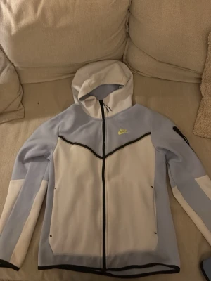 Nike Tech Fleece hoodie ljusblå/vit - Nike Tech Fleece hoodie i ljusblått och vitt med svarta detaljer. Dragkedja framtill, huva och två sidofickor. Ikonisk Nike-logga i neongult på bröstet. Tillverkad i mjukt och lätt material som passar perfekt för en chill och sportig look.