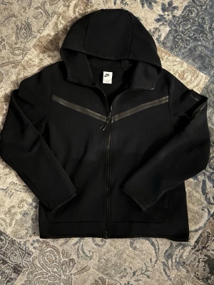 Svart hoodie från Nike med dragkedja - Svart hoodie från Nike med hel dragkedja framtill och två snedställda reflexdetaljer över bröstet. Klassisk huva och Nike-logga på vänster sida. Tillverkad i ett mjukt och bekvämt material, perfekt för en sportig och avslappnad stil.