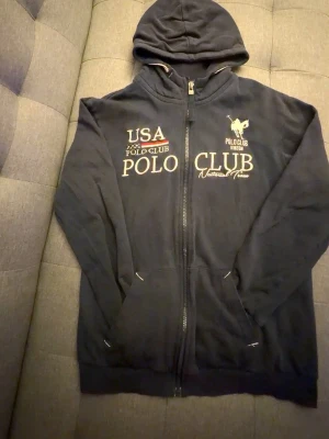 Marinblå hoodie från Polo Club - Mörkblå hoodie med dragkedja från Polo Club. Tryck på bröstet med texten 'USA Polo Club' och en hästlogga. Utrustad med huva och två fickor framtill. Perfekt för en avslappnad och sportig stil.