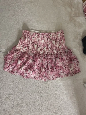 Blommig volangkjol från Gina Tricot - Supersöt kort kjol från Gina Tricot med blommigt mönster i rosa och vitt. Kjolen har smockad midja och volang nertill, perfekt för en trendig och feminin look. Lätt och luftig känsla.
