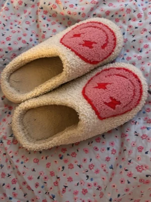 Beiga tofflor med rosa smiley - Mysiga beiga tofflor i fluffigt material med rosa smiley-mönster på ovansidan. Tofflorna har rund tå och platt sula, perfekta för att chilla hemma. De är fodrade för extra komfort och har en avslappnad look.
