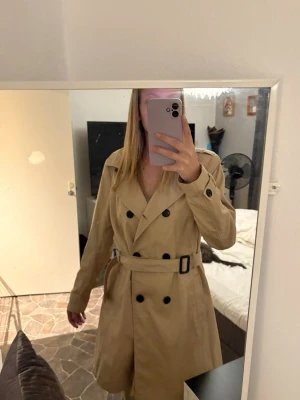 Trenchcoat - Stilren beige trenchcoat med dubbelknäppning, bälte i midjan och klassiska svarta knappar. Jackan har långa ärmar med knäppning vid ärmslut och en snygg krage. Perfekt för dig som vill ha en tidlös och trendig look.