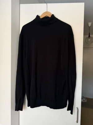 Svart polotröja i stickat material - Klassisk svart stickad polotröja med lång ärm och ribbade muddar. Tröjan har en hög polokrage och är tillverkad i ett mjukt, tunt material som passar perfekt till lager-på-lager. Enkel och stilren design utan synliga loggor eller mönster.