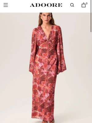 Röd blommig långklänning från Adoore - Säljer en röd långklänning från Adoore med blommigt mönster. Klänningen har v-ringning, långa ärmar och knappar framtill. Perfekt för dig som vill sticka ut med en färgstark och elegant look. En knapp har lossnat, syns om man zomar in på bild. Inget jag la märke till förens jag kollade noga alltså ingen som syns när man bär den.