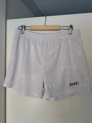 Vita sportiga shorts från JAKO - Säljer ett par vita shorts från JAKO med diskret mönster och elastisk midja. Shortsen har en avslappnad passform och är perfekta för sommarens alla aktiviteter. Materialet känns lätt och svalt, och loggan är broderad på ena benet.