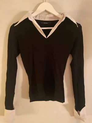 Svart topp med vit krage och manschetter - Stilren svart topp från Vero Moda med vit klassisk skjortkrage och vita manschetter. V-ringad design som ger en snygg kontrast mellan svart och vitt. Figurnära passform och långa ärmar. Perfekt för en trendig och chic look.