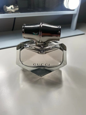 Gucci bamboo  - Snygg parfymflaska från Gucci med en unik, geometrisk form och ett stort silverfärgat lock. Flaskan är genomskinlig med svart text och känns riktigt lyxig och modern. Perfekt för dig som gillar exklusiva dofter och vill ha en stilren accessoar på sminkbordet.