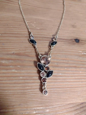 Pilgrim guldigt halsband med svarta och bruna stenar - Supersnyggt guldfärgat halsband med metallkedja och dekorativa svarta och bruna stenar/kristaller i olika storlekar. Känner inte till vad det är för material. 35 + 8 cm långt. Danish Design. I fint använt skick. Kan skickas med 1 frimärke (22 kr).