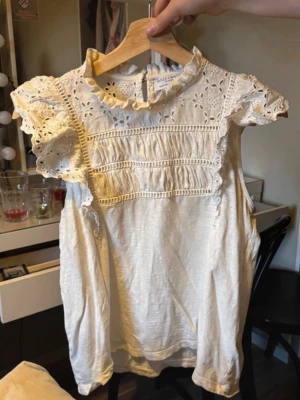 Vit blus med spets och volang - Söt vit blus från Holly & Whyte med broderade spetsdetaljer och volang vid axlarna. Blusen har rund hals med liten volangkant och korta ärmar. I storlek S