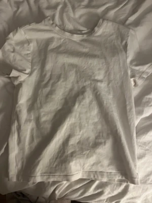 Vit basic t-shirt i bomull - En enkel vit t-shirt i mjuk bomull. Klassisk rund halsringning och korta ärmar. Perfekt att matcha med jeans eller shorts för en clean och stilren look.