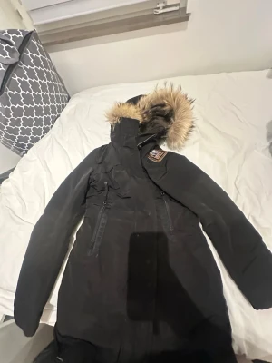 Svart parkas med fuskpäls från Didriksons - Snygg svart parkas från Didriksons med huva och fuskpälsdetalj. Jackan har dragkedja framtill, två stora fickor och en patch på bröstet. Perfekt för kalla dagar med sin långa modell och varma material. Äkta pjs! Päls äkta!