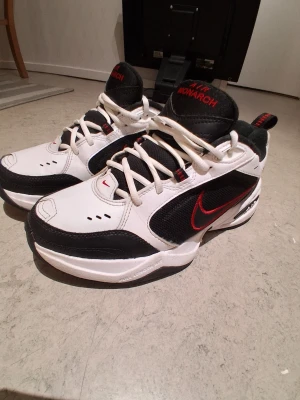 Nike Air max - Nike Air Monarch sneakers i vitt och svart med röda detaljer. Skorna har en klassisk chunky siluett, snörning och en tydlig röd swoosh på sidan. Ovandelen är i skinn och syntet med meshpaneler för extra andningsförmåga. Perfekta för en sportig och retro look.