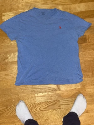 Blå t-shirt från Polo Ralph Lauren - Säljer en blå t-shirt från Polo Ralph Lauren med klassisk röd logga broderad på bröstet. T-shirten har rund halsringning och korta ärmar. Tillverkad i mjuk bomull som känns skön mot huden. Perfekt för en avslappnad och stilren look.