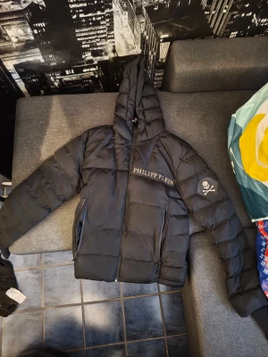 Svart pufferjacka  - Säljer en svart pufferjacka  med huva och dragkedja. Jackan har logotypen tryckt på bröstet och ryggen samt en döskalle-detalj på ena ärmen. Perfekt för dig som vill ha en streetstyle-look med exklusiv känsla.