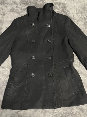 Svart skepparkavaj med hög krage - Stilren svart skepparkavaj med dubbelknäppning och hög krage. Jackan har långa ärmar, två fickor framtill och markerade sömmar för en snygg siluett. Perfekt för dig som sopar gatan med din stil.