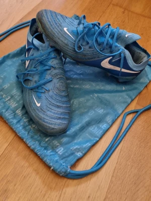 Blå Nike fotbollsskor med snörning - Säljer ett par blå Nike fotbollsskor med vit swoosh och snörning. Skorna har en rund tå och platt sula med dobbar för bra grepp på planen. Tillverkade i syntetmaterial och designade för att ge bra bollkontroll och komfort under matchen.