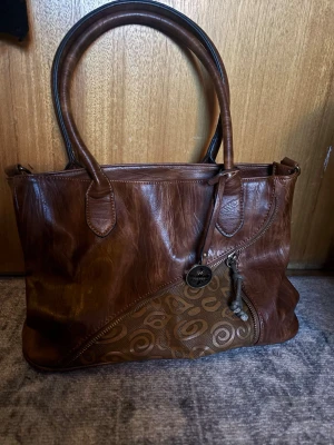 Brun handväska från Esprit i skinn - Snygg brun handväska från Rieker i skinn med dubbla handtag och dekorativt mönstrad panel framtill. Väskan har en dragkedja baktill och ett hängande Rieker-märke. 