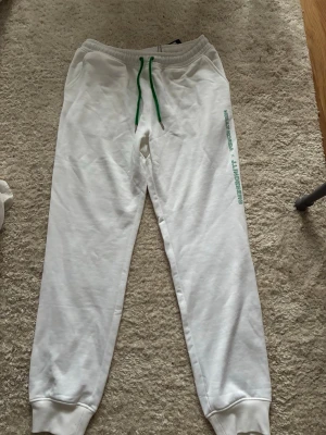 Vita mjukisbyxor med grön snörning - Snygga vita mjukisbyxor från Nella Korda x J.Lindeberg med grön snörning i midjan och tryckt logga längs benet. Byxorna har resår i midjan och vid bensluten. Orginalpris var 800kr. (Skriv om ni har frågor💕)