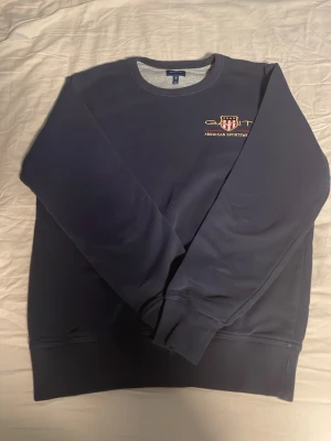 Mörkblå sweatshirt från GANT - Mörkblå sweatshirt från GANT med rund halsringning och broderad logga samt texten 'American Sportswear' på bröstet. Tröjan har ribbade muddar vid ärmslut och nederkant. Perfekt för en avslappnad och sportig stil.