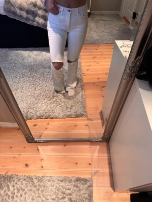 Vita ripped jeans med hög midja - Säljer ett par vita jeans med hög midja och raka ben. Jeansen har snygga slitningar över knäna och klassisk femficksdesign. Perfekta för dig som vill ha en clean men edgy look. Materialet är mjukt och stretchigt jeans.