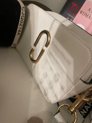 Vit axelväska från Marc Jacobs - Snygg vit axelväska från Marc Jacobs med guldiga metalldetaljer och bred, randig axelrem med logotyp. Väskan har en stilren, rektangulär form och är tillverkad i skinn. Perfekt för att lyfta din outfit med en trendig touch.