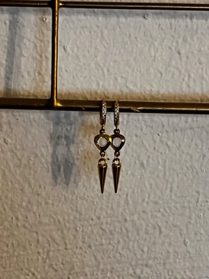 Guldpläterade örhängen med strass och spets - Säljer ett par trendiga guldpläterade örhängen med små gnistrande stenar på ringen och en hängande klar sten i droppform. Nedanför stenen hänger en spetsig kon i metall som ger en edgy look. Perfekta för dig som gillar statement-smycken.
