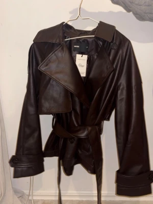 Brun skinnjacka med bälte från Bik Bok - Superfin mörkbrun skinnjacka från Bik Bok i trendig modell med breda slag, bälte i midjan och dekorativa detaljer vid ärmsluten. Jackan har en klassisk trenchcoat-look och är gjord i ett glansigt skinnmaterial❣️passar s-m, endast testad