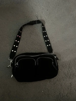 Svart axelväska med nitar från Gina Tricot - Cool svart axelväska från Gina Tricot med bred axelrem dekorerad med silverfärgade och rosa nitar. Väskan har två framfickor med dragkedja och ett stort huvudfack. Perfekt för dig som gillar en edgy stil och vill ha plats för allt viktigt.