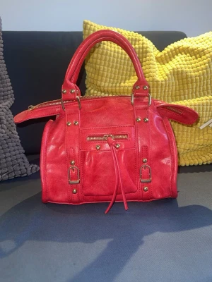 Röd handväska från Italien - Snygg röd handväska i skinnimitation med guldfärgade nitar och spännen. Väskan har dubbla handtag, dragkedja upptill och en liten ficka framtill med dragkedja och kommer med tillhörande axelrem. 20x22x12 i storleken. ✨💕 Nypris 350kr men säljer för 175kr